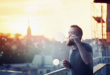 Modern happy young man with a beard fun Vaporizers, smoke and bubbles on the terrace. In the background, the evening sunset over the old city