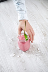 Top view close focus hand takes rustic jar with cold fresh berry smoothie isolated on wooden table with golden straw inside surrounded with crashed melted ice cubes Summer healthy resfreshment drink