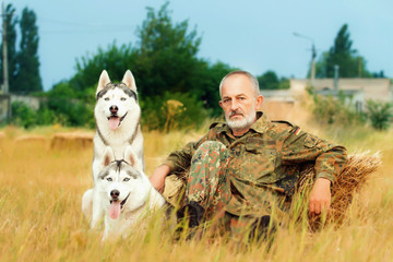 Old man with a beard sitting on a haystack with their dogs, enjoying summer sunset. Siberian Husky in the countryside. Harmony between man and nature. Hunter with dogs in the field.