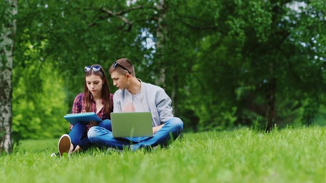 Young man and woman working together on a laptop and a tablet. Sitting on the grass in the park