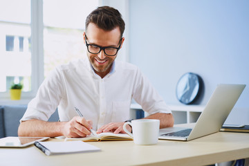 Happy man working from home office doing notes sitting at desk
