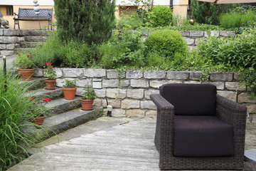 Fototapeta premium Vintage Gestaltung eines Hanggrundstücks: Garten mit Terrasse aus Holz und Treppe und Mauer aus Naturstein, dekoriert mit Grünpflanzen und Gräsern, Gartenmöbel Sessel aus Rattan 