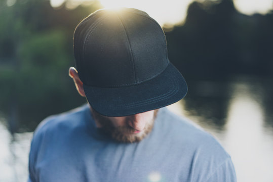 Photo Bearded Young Man Wearing Black Blank Cap. Green City Park Lake Background and Sunset effect. Front view. Horizontal Mockup