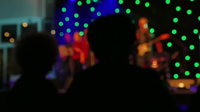 Two silhouettes slowly enjoy the concert, in the background musicians, perform the concert