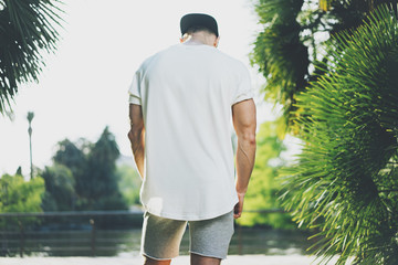 Photo Bearded Muscular Man Wearing White Blank t-shirt and shorts in summer time. Green City Garden Park Background. Back view. Horizontal Mockup