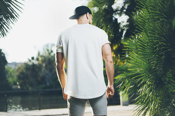 Photo Bearded Muscular Man Wearing White Blank t-shirt, snapback cap and shorts in summer time. Green City Garden Park Background. Back view. Horizontal Mockup