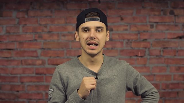 Young man in black cap, pronounce words in camera, hold mini microphone on pullover. Brick wall on background. Blogger