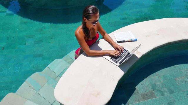 Woman working on her laptop computer sitting at poolside. Beautiful young female model working freelance at swimming pool on laptop.