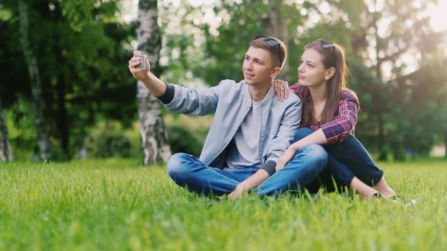 Young couple photographing themselves on the phone, making funny faces. Sitting in the park on the grass