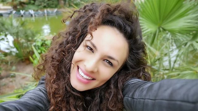 Portrait of happy young woman with beautiful curly hair taking selfie in the park, slow motion