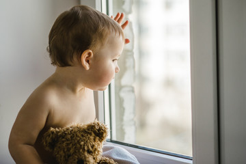 a little boy sitting by the window with a teddy bear in his hands