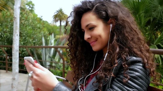 Young woman with beautiful curly hair listening to music in the park, slow motion