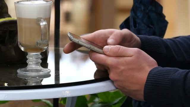 Male hands using smartphone and having cappuccino at cafe in shopping mall. Close-up of man browsing information and scrolling pictures on smart phone