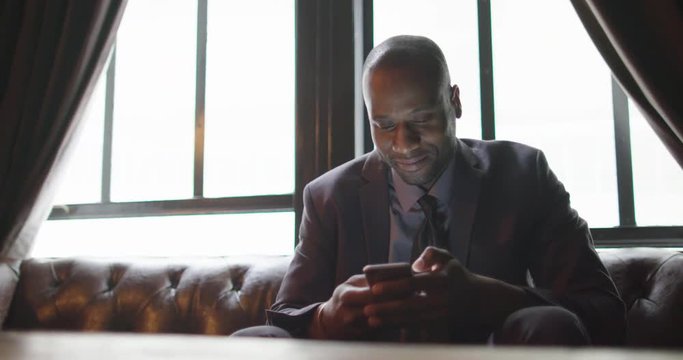 African American business man sitting on a leather sofa and using a cellphone in a relaxed indoor setting.  Slow motion mid shot recorded at 60fps.