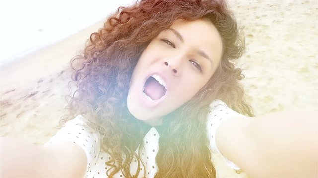 Portrait of happy young woman with beautiful curly hair taking selfie on the beach, slow motion