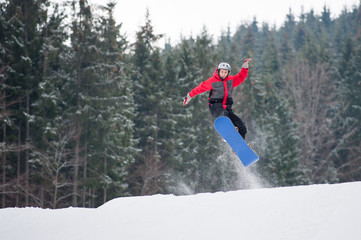 Naklejka na ściany i meble Young male on the snowboard jumping over the slope in winter with snowy slope and snow-covered firs in background, extreme sport