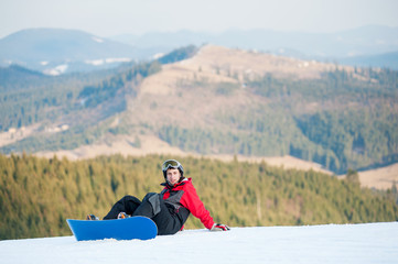 Guy with snowboard wearing helmet, red jacket, gloves and pants sitting on snowy slope on top of a mountain and looking to the camera, with an astonishing view on mountains, hills, forest