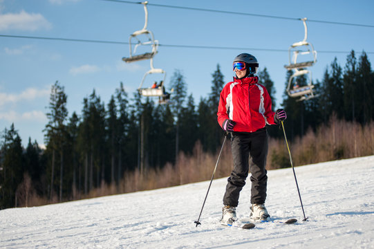 Woman skier wearing helmet, red jacket and ski goggles standing with skis on snowy slope and looking away in sunny evening with forest and blue sky in background.