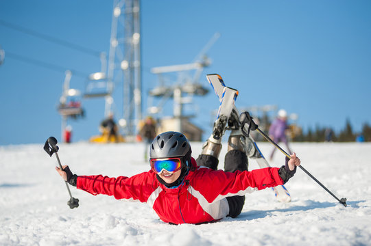 Skier female in ski goggles lying with raised arms on snowy slope at mountain top in sunny day with ski lifts and blue sky in background. Close-up