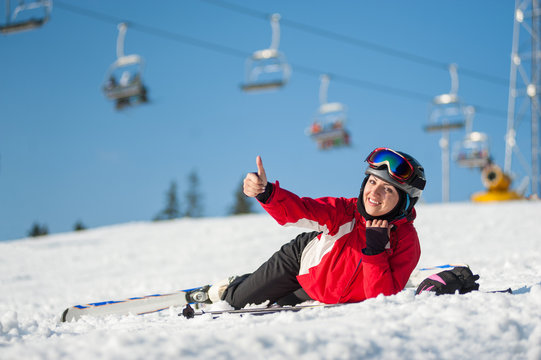 Smiling female skier lying with skis on snowy at mountain top, showing thumb up gesture of good class and looking at the camera in sunny day at winter resort with ski lifts and blue sky in background