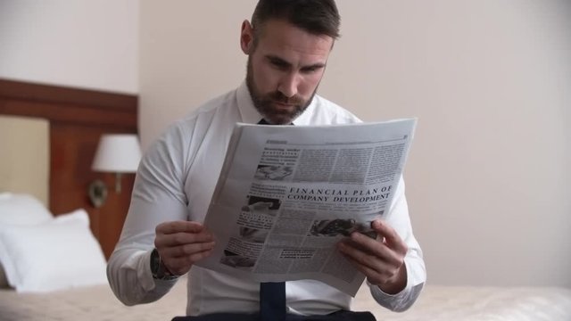 Serious businessman in shirt and tie sitting in his hotel room and reading financial article in newspaper 