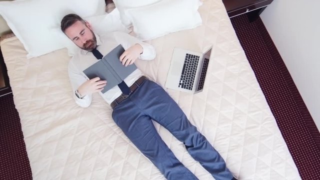 High angle view of businessman lying on the bed and reading book then he closing book and falling asleep. There is laptop on the bed and smart phone on bedside table