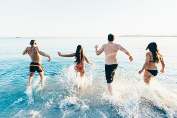 Rear view of a group of young multiethnic friends women and men running on the beach to the sea in summertime - happiness, summer, friendship concept