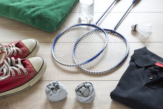 Rackets for badminton, shuttlecock, polo shirts, shoes, towel and water on a wooden floor.