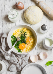 Raw ingredients for baking a pie with spinach - eggs, cheese, dough, spinach, cream on light wooden background. Baking rustic background
