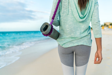 Fitness woman carrying yoga mat with strap on beach going to class training. Closeup of sports equipment, back view of fit athlete in activewear showing fashion leggings and turquoise hoodie. - obrazy, fototapety, plakaty
