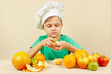 Young boy in chefs hat peeling fresh orange at the table with fruits