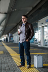 Young regular man standing waiting near the airport with cup of coffee and silver case