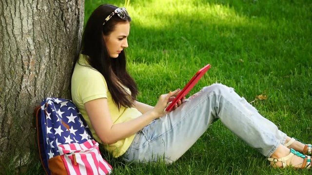footage young beautiful woman sitting near a tree on the grass in the park and use the app in your comuter tablet