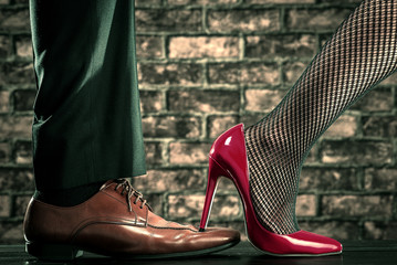赤いハイヒールの女性が男性の革靴を踏んでいる - obrazy, fototapety, plakaty