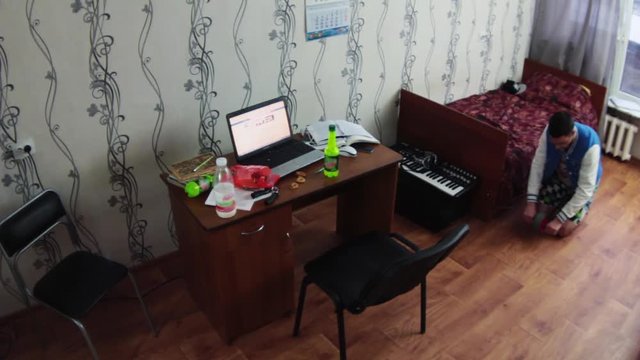 Young boy exercise with sport disk on floor in dormitory room. Table with notebook, bottles, cookies. Student. 