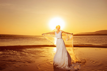 Fototapeta na wymiar Beautiful bride in long wedding dress is holding veil at sea sunrise