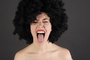 Funny woman in a wig is shouting with tongue out. Studio shot with dark background.