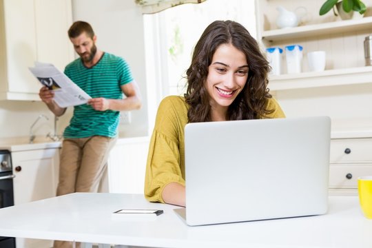 Woman using laptop while man reading newspaper in background