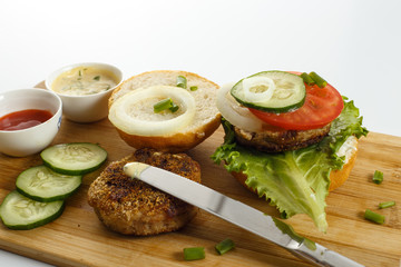 Plakat Cooking process of a sandwich burger, ingredients on wooden cutting board on wooden table against white background, fresh vegetables, herbs, fried meat, buns, sauces and knife