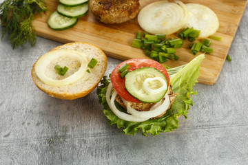 Cooking process of a sandwich burger, ingredients on wooden cutting board on wooden table against white background, fresh vegetables, herbs, fried meat, buns, sauces and knife