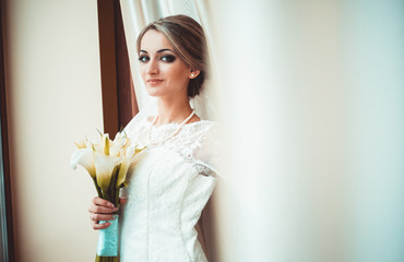 Close up portrait of happy stylish blond bride under the veil in luxury white dress standing near the window