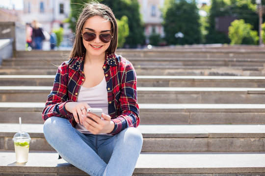 Happy young woman with a fresh cup sitting on the stairs and using her smartphone for communication.