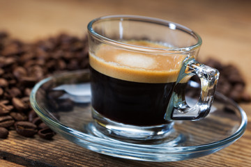 A transparent glass cup of hot espresso on a saucer standing on a wooden table top, surrounded by coffee beans
