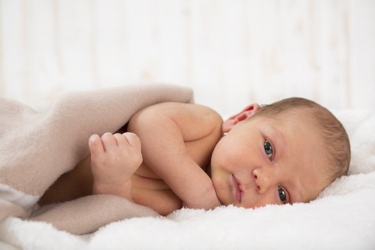 Newborn baby lying on a white blanket underneath a tan colour blanket looking at the camera