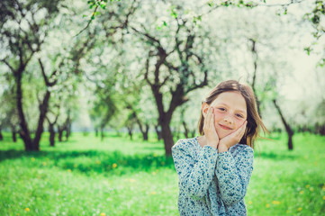 Fototapeta na wymiar Smiling little girl in the garden, holding hands your face. Child in spring fruit orchard.
