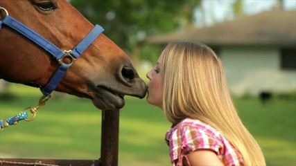 A pretty country girl gets an appreciative kiss from her horse who she is feeding a carrot. Camera tracks action