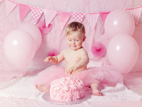 Portrait of cute adorable Caucasian baby girl with blue eyes in pink tutu skirt celebrating her first birthday with gourmet cake and balloons looking away, cake smash first year concept