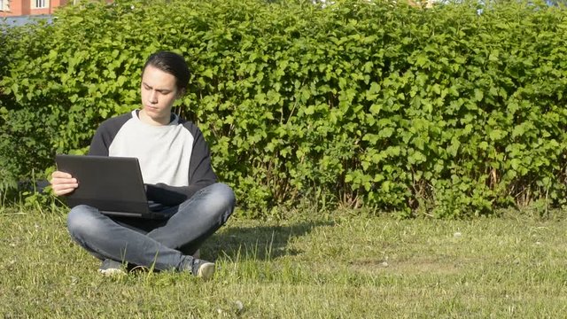 Handsome young man sitting on the grass in the park and to work with laptop.