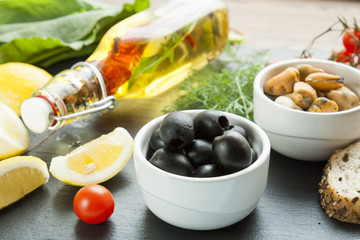 Black olives and mussels in ceramic bowls, fresh tomatoes, sliced lemon, bread and bottle of olive oil with spices on wooden board. Mediterranean food on black stone board