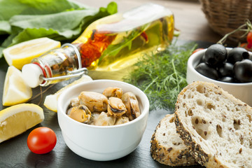 Mussels and black olives and in ceramic bowls, fresh tomatoes, sliced lemon, bread and bottle of olive oil with spices on black stone board. Mediterranean food on black stone board